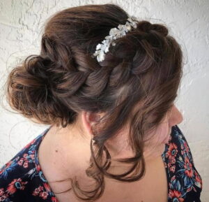 Woman with brown hair styled in a braided updo with loose curls, wearing a silver floral hair accessory and a floral-patterned dress, facing to the side. - Escape Salon | Nokomis, FL