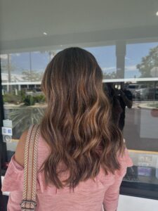 A person with wavy, shoulder-length brown hair stands facing away from the camera, wearing a pink off-the-shoulder top and a patterned strap purse. - Escape Salon | Nokomis, FL