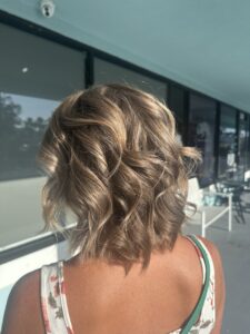 A person with short, wavy, highlighted blonde hair stands outside in sunlight, wearing a sleeveless patterned top with thin straps. - Escape Salon | Nokomis, FL