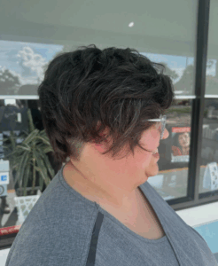 A person with short, dark, wavy hair and glasses, wearing a gray shirt, is pictured in profile outside a building with large windows. - Escape Salon | Nokomis, FL