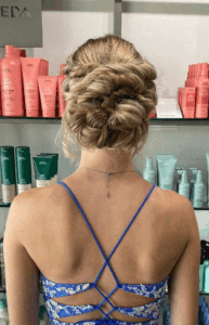 A woman with blonde hair styled in an intricate updo stands in front of shelves stocked with hair care products, wearing a blue strappy dress. - Escape Salon | Nokomis, FL