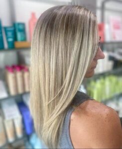 A person with straight, shoulder-length blonde hair stands in a store or salon, facing away from the camera. Shelves with hair products are visible in the background. - Escape Salon | Nokomis, FL