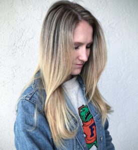 A woman with long, straight blonde hair looks down, wearing a denim jacket over a shirt with a cartoon alligator graphic on it, standing against a white textured wall. - Escape Salon | Nokomis, FL