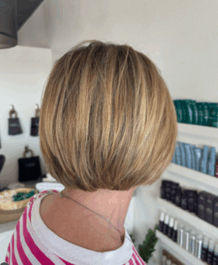 A person with short, straight blonde hair is shown from the back in a salon setting, wearing a pink and white striped shirt. - Escape Salon | Nokomis, FL