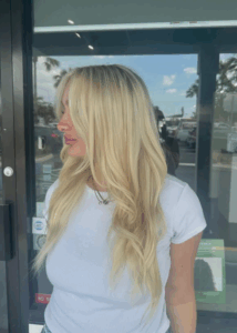 A woman with long, wavy blonde hair wearing a white short-sleeve top stands outside in front of a glass door, looking to her left. - Escape Salon | Nokomis, FL