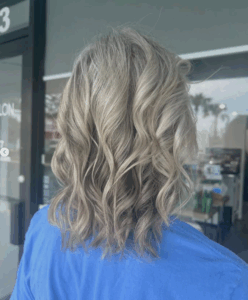 Person with shoulder-length, wavy, gray-blonde hair wearing a blue top, standing indoors facing away from the camera. - Escape Salon | Nokomis, FL