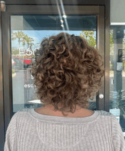 Person with short, curly brown hair stands facing away from the camera, wearing a light gray textured sweater, in front of a glass door with reflections and signage. - Escape Salon | Nokomis, FL