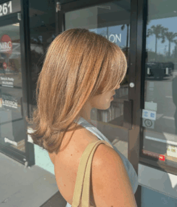 A person with shoulder-length, layered light brown hair stands outside a glass storefront in sunlight, holding a beige bag on their shoulder. - Escape Salon | Nokomis, FL