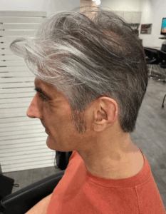 A man with short, neatly styled salt-and-pepper hair sits in a salon chair, wearing an orange shirt. - Escape Salon | Nokomis, FL