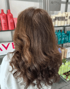 Person with long, wavy brown hair stands in front of shelves displaying various Aveda hair products in bottles of different colors. - Escape Salon | Nokomis, FL