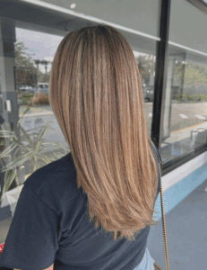 A person with straight, shoulder-length, highlighted hair stands outside near a glass window and plants, facing away from the camera. - Escape Salon | Nokomis, FL