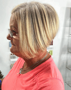 A woman with short, straight, highlighted blonde hair styled in a bob, wearing glasses and a pink top, is pictured in profile against a light background. - Escape Salon | Nokomis, FL