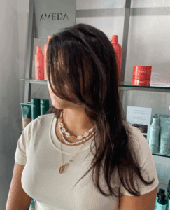 A woman with long dark hair partially covering her face stands in front of salon shelves with various hair products and an AVEDA sign. - Escape Salon | Nokomis, FL