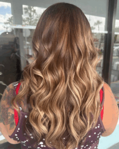 A person with long, wavy, light brown and blonde balayage hair stands facing away from the camera, wearing a sleeveless polka dot top. - Escape Salon | Nokomis, FL