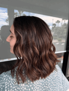A person with shoulder-length, wavy brown hair with subtle highlights is shown in profile, standing in front of a window. - Escape Salon | Nokomis, FL