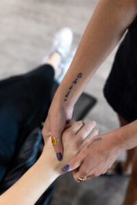 Two hands are clasped; one has a gold ring and a small heart tattoo on the forearm. The person in the background is wearing black pants and white shoes. - Escape Salon | Nokomis, FL