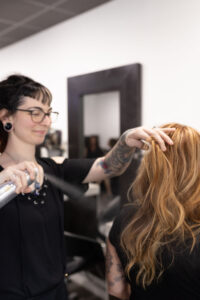 A hairstylist sprays hair product onto a client's wavy, light brown hair in a salon. - Escape Salon | Nokomis, FL